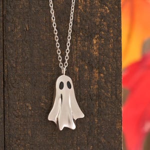 May include: A silver-colored ghost pendant necklace. The ghost has two black oval eyes and a flowing body. The necklace chain is also silver and hangs against a dark brown wooden background. The background has blurred orange and red colors.