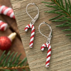 May include: A pair of silver leverback earrings with candy cane charms. Each charm is a red and white striped candy cane. The earrings are displayed on a wooden surface with Christmas decorations in the background.