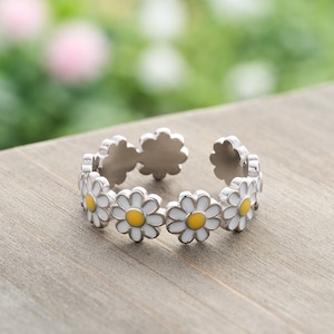 May include: A silver-colored ring featuring a daisy design. Each daisy has white petals and a yellow center. The ring is open, allowing for size adjustment. The background is a wooden surface with a blurred green and pink backdrop.