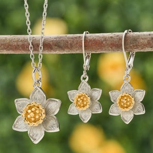 May include: A silver-toned necklace pendant and matching earrings, each featuring a daffodil design with a gold-colored center. The jewelry is displayed on a natural branch against a blurred green background, showcasing a floral theme.