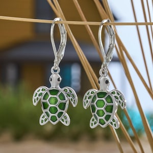 May include: A pair of silver-toned leverback earrings featuring a turtle design. The turtles have a green, honeycomb-patterned shell and detailed flipper design. The earrings are displayed against a blurred background of greenery and dry grass.