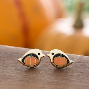 May include: A pair of gold-tone earrings featuring a small bird design with an orange pumpkin inside. The earrings are on a wooden surface.