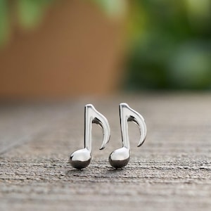 May include: A pair of silver-colored musical note earrings. The earrings are shaped like eighth notes, with a rounded bottom and a curved stem. They are displayed on a textured, wooden surface.