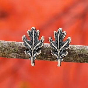 May include: A pair of leaf-shaped stud earrings. Each earring features a detailed oak leaf design with dark gray and silver tones. The earrings are displayed on a small branch against a blurred red background.