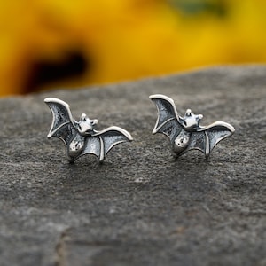 May include: A pair of silver-toned bat stud earrings. Each earring features a detailed bat design with outstretched wings. The bats have a textured surface and are set against a blurred yellow and gray background.