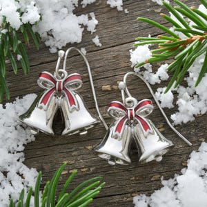 May include: A pair of silver-toned drop earrings featuring two small bells tied together with a red and white enamel bow. The earrings have curved silver ear wires and a polished metallic finish, designed to hang as festive holiday jewelry.