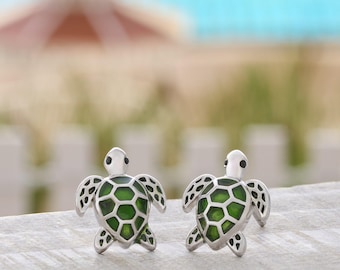 May include: A pair of silver-toned stud earrings shaped like sea turtles. Each turtle features a vibrant green enamel shell with a detailed black geometric pattern and small black eyes, crafted with a polished metallic finish on the head and limbs.