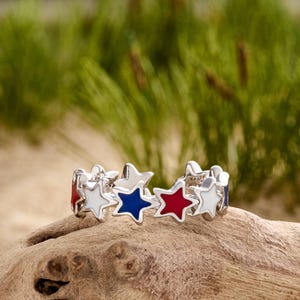 May include: A silver ring with a patriotic theme, featuring alternating red, white, and blue star-shaped enamel designs. The ring is displayed on a piece of driftwood, with a blurred green background.