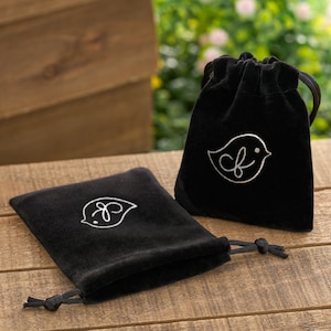 May include: Two black velvet drawstring pouches with a white embroidered bird logo. One pouch is open, revealing the drawstring, while the other is closed. The pouches are on a wooden surface, with a blurred background of greenery.