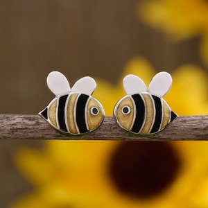 May include: A pair of bee-shaped earrings. Each earring features a yellow body with black stripes, white wings, and small black eyes. The earrings are set against a blurred background of a sunflower.