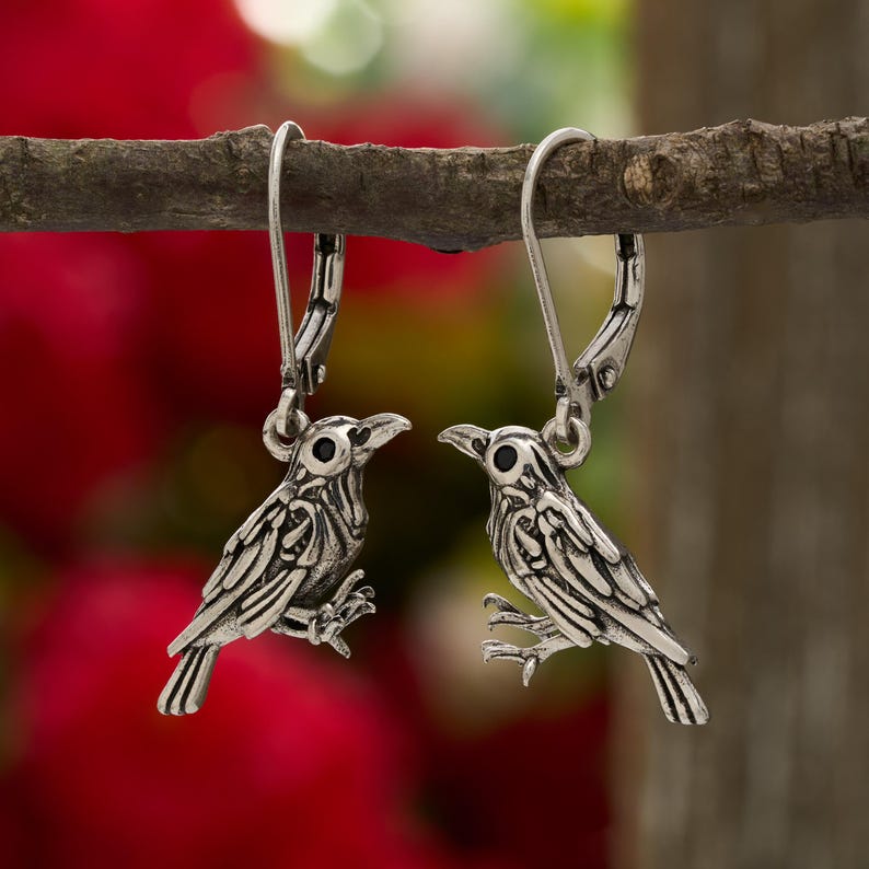 May include: A pair of silver-colored bird earrings. Each earring features a detailed bird design with a textured surface, black eyes, and a hinged ear wire. The earrings are displayed on a small branch against a blurred red and green background.