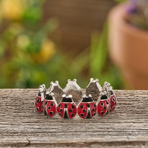 May include: A silver-toned ring featuring a continuous band of small, red and black enameled ladybug charms. Each ladybug is detailed with black spots and a central line, creating a repeating pattern around the entire circumference of the ring.