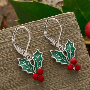 May include: A pair of silver-tone leverback earrings. Each earring features a green holly leaf design with white accents and three red berries. The earrings are set against a wooden surface.