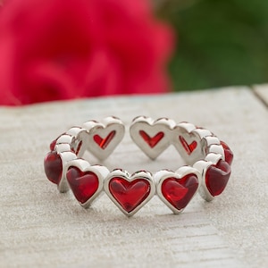 May include: A silver-toned ring featuring a repeating heart design. Each heart is outlined in silver and filled with a vibrant red material, creating a romantic and eye-catching piece of jewelry.