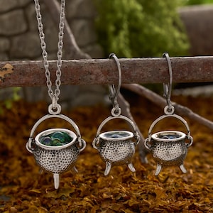 May include: A silver-toned jewelry set consisting of a necklace and a pair of earrings. Each piece features a miniature three-legged cauldron charm with a textured surface and a small handle, containing tiny, translucent green and blue spheres.