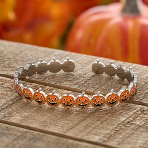 May include: Silver-toned cuff bracelet featuring a repeating pattern of orange jack-o'-lantern faces with black smiles and eyes. The bracelet is open-ended and rests on a wooden surface, with pumpkins in the background.
