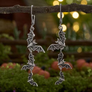 May include: A pair of silver-toned drop earrings featuring a vertical chain of three bat-shaped charms. Each earring is suspended from a lever-back closure, showcasing detailed wings and pointed ears on the bat figures with an antiqued finish.