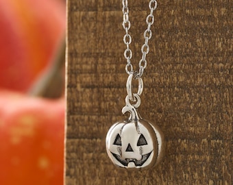 May include: A silver-colored necklace with a pumpkin pendant. The pendant has a carved jack-o'-lantern face with triangular eyes and a smiling mouth. The necklace chain is delicate and hangs against a wooden background. The pumpkin pendant is approximately 0.75 inches in diameter.
