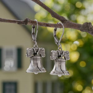 May include: A pair of silver-toned earrings shaped like the Liberty Bell. The earrings are suspended from lever-back closures and are hanging from a tree branch. The background is blurred, with a house and green foliage visible.