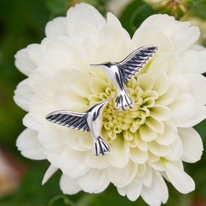 May include: Two silver hummingbird earrings on a white flower. The hummingbirds are facing opposite directions and have detailed wings.