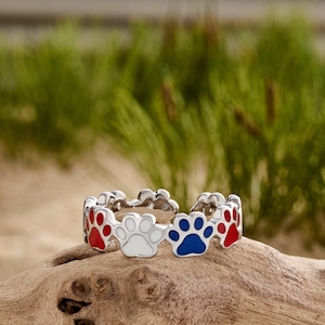 May include: A silver ring featuring a repeating pattern of paw prints in red, white, and blue enamel. The ring is displayed on a piece of driftwood, with a blurred background of green grass.