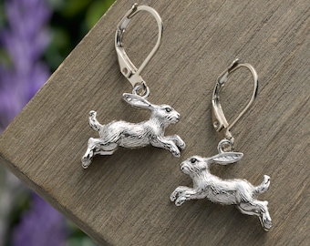 May include: A pair of silver-toned leverback earrings. Each earring features a detailed, running rabbit charm. The earrings are displayed on a wooden surface with a blurred background of purple flowers.
