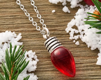 May include: A silver chain necklace with a red Christmas light bulb pendant. The bulb has a silver base with black stripes. The necklace is on a wooden surface with artificial snow and pine needles.