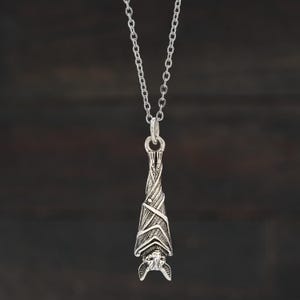 May include: A silver-colored bat pendant necklace. The bat is hanging upside down, with detailed wings and a textured body. The chain is a delicate silver link. The pendant is suspended from a small loop, and the background is a dark, blurred wood grain.