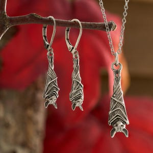 May include: A silver-toned jewelry set featuring a bat design. The set includes a pair of dangle earrings and a pendant necklace. The bats are depicted hanging upside down, with detailed wings and faces. The jewelry hangs from a branch against a red background.