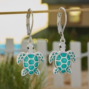 May include: A pair of silver-toned leverback earrings featuring sea turtle charms. Each turtle has a white head with black eyes and a shell filled with vibrant turquoise enamel, showcasing a detailed hexagonal pattern on the back of the shell.