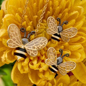 May include: A gold-toned bee pendant necklace and matching bee stud earrings. Each bee features black and gold striped bodies, dark metallic heads, and translucent wings with intricate gold vein detailing, resting on a bright yellow flower.