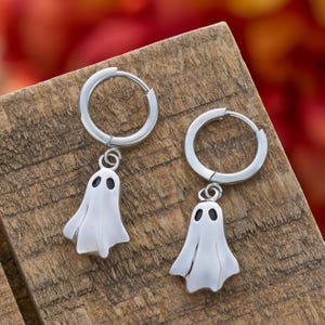 May include: A pair of silver-colored hoop earrings with a dangling ghost charm. Each ghost charm has two black oval eyes. The earrings are displayed on a wooden surface with a blurred red and orange background.