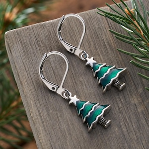 May include: A pair of silver-toned leverback earrings. Each earring features a green and white enamel Christmas tree design with a star at the top. The earrings are displayed on a wooden surface with a sprig of pine needles.