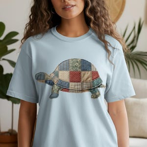 May include: Light blue t-shirt featuring a patchwork turtle design. The turtle is made of various fabric patterns in shades of blue, red, and green. The shirt has short sleeves and a crew neck, perfect for casual wear.