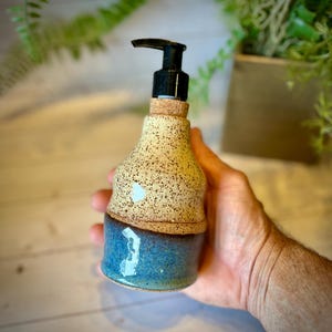 May include: A ceramic soap dispenser featuring a speckled cream-colored top section and a smooth, glossy blue bottom section. The dispenser is topped with a black plastic pump mechanism and a small cork collar at the neck of the bottle.
