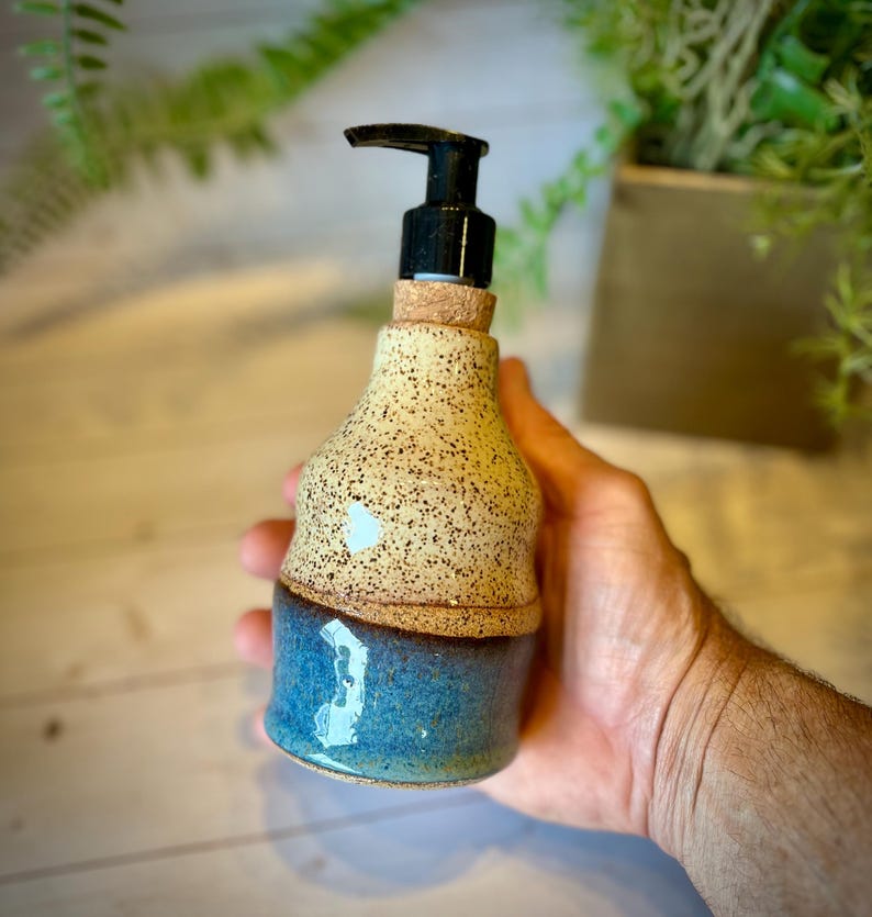 May include: A ceramic soap dispenser featuring a speckled cream-colored top section and a smooth, glossy blue bottom section. The dispenser is topped with a black plastic pump mechanism and a small cork collar at the neck of the bottle.