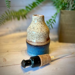 May include: A ceramic bottle with a speckled cream-colored top section and a solid blue bottom section. The bottle has a rounded, tapered neck and a wide base. A separate black pump dispenser with a cork stopper is positioned in front of the bottle.