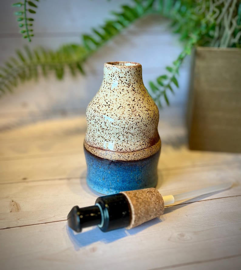 May include: A ceramic bottle with a speckled cream-colored top section and a solid blue bottom section. The bottle has a rounded, tapered neck and a wide base. A separate black pump dispenser with a cork stopper is positioned in front of the bottle.