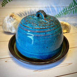 May include: A ceramic butter dish with a dome-shaped lid featuring a vibrant blue glaze and a textured brown rim. The lid has a small handle at the top, and the base is a matching dark brown plate with a smooth, glossy finish and blue accents.