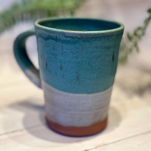 May include: Handmade ceramic mug with a teal blue upper section, a white middle, and a reddish-brown base. The mug has a curved handle and a textured surface. A unique piece for enjoying coffee or tea.