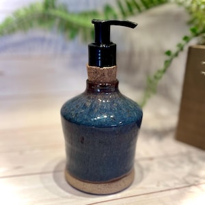 May include: A ceramic soap dispenser with a glossy, mottled blue glaze on the upper body and a natural, unglazed beige base. The neck features a cork collar and a black plastic pump mechanism for dispensing liquid soap or lotion.