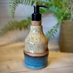 May include: A ceramic soap dispenser with a speckled cream-colored top section and a solid blue bottom section. The vessel features a cork stopper at the neck and a black plastic pump mechanism at the top for dispensing liquid soap or lotion.