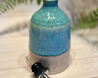 May include: A ceramic oil cruet featuring a two-tone glaze with a speckled turquoise top and a speckled cream-colored base. The bottle has a narrow neck and includes a removable stainless steel pourer spout with a black rubber stopper and tip.
