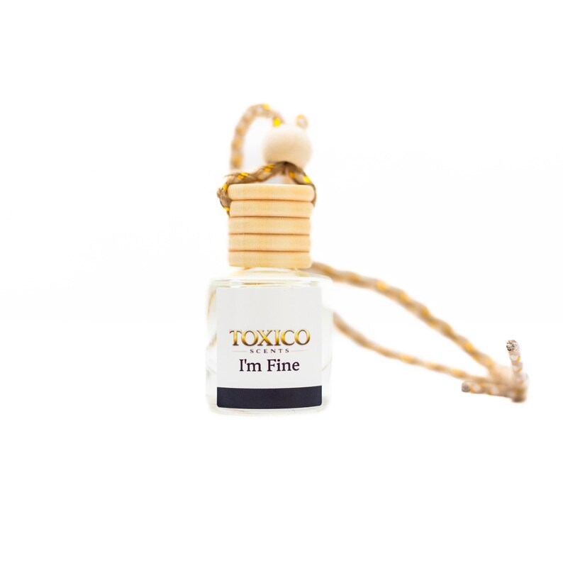 Puede incluir: Una peque&ntilde;a botella de vidrio transparente con tapa de madera y cord&oacute;n tejido para colgar. La botella tiene una etiqueta con el texto "TOXICO SCENTS" y "I'm Fine". Dise&ntilde;ada para contener fragancias o aceites esenciales.