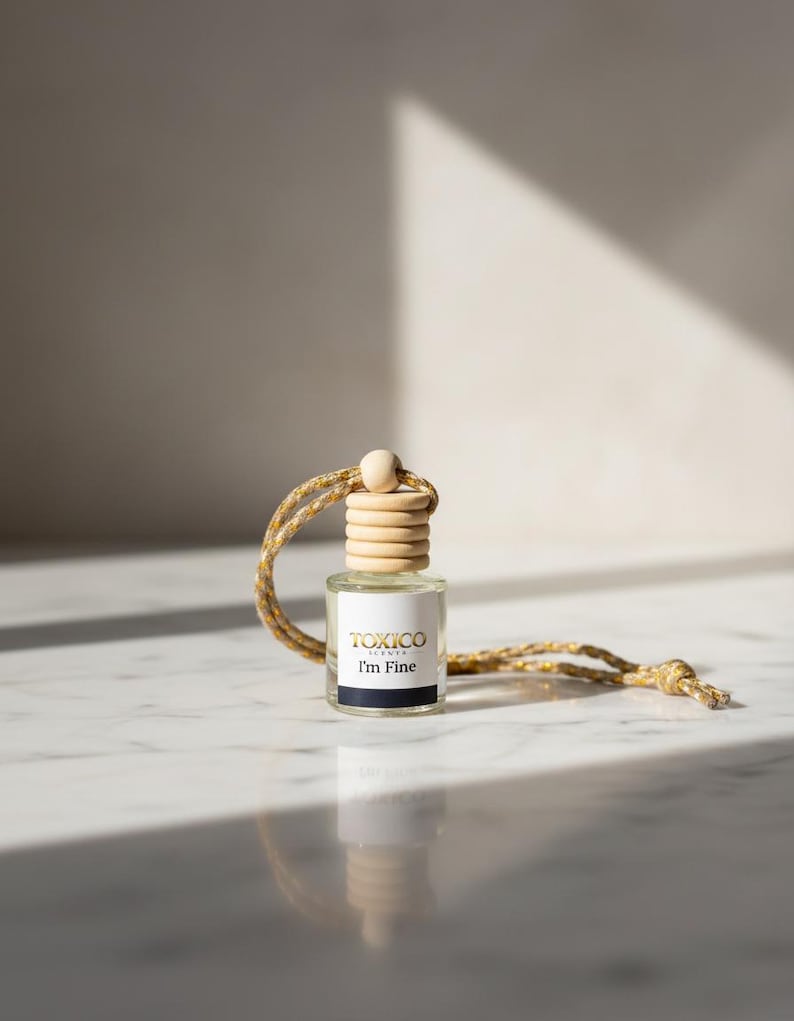 Puede incluir: Peque&ntilde;a botella de vidrio transparente con tap&oacute;n de madera y cuerda dorada y beige. La etiqueta dice "TOXICO I'm Fine" en negro y dorado. La botella est&aacute; dise&ntilde;ada para colgar, probablemente un ambientador o fragancia.