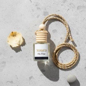 Puede incluir: Peque&ntilde;a botella cuadrada de vidrio con tap&oacute;n de madera y cord&oacute;n trenzado, etiquetada "TOXICO SCENTS I'm Fine". El cord&oacute;n es dorado y beige. Tambi&eacute;n hay un p&eacute;talo seco y una peque&ntilde;a piedra gris.