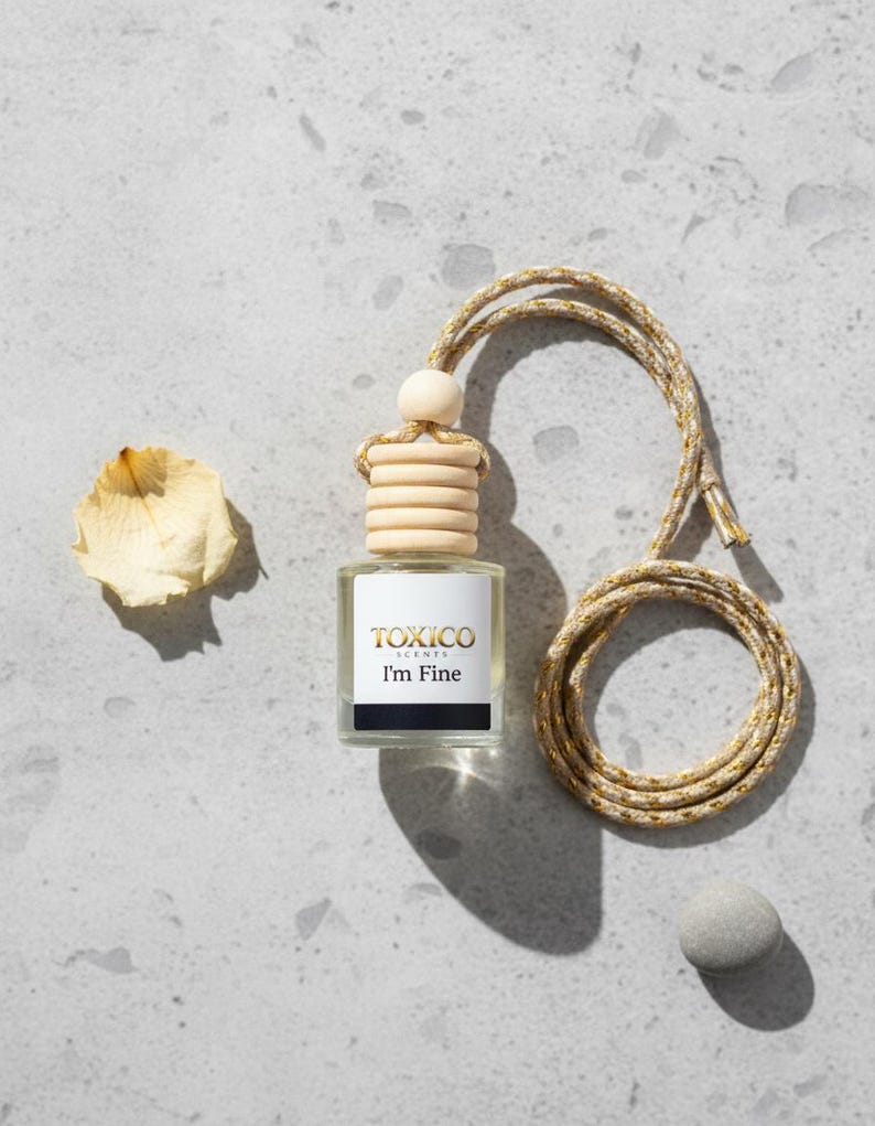 Puede incluir: Peque&ntilde;a botella cuadrada de vidrio con tap&oacute;n de madera y cord&oacute;n trenzado, etiquetada "TOXICO SCENTS I'm Fine". El cord&oacute;n es dorado y beige. Tambi&eacute;n hay un p&eacute;talo seco y una peque&ntilde;a piedra gris.