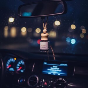 Puede incluir: Un ambientador de coche con el texto "TOXICO I'm Fine" cuelga del espejo retrovisor. Se ve el interior del coche, con el salpicadero, los indicadores y una pantalla iluminada en azul. El fondo muestra luces borrosas de la ciudad.