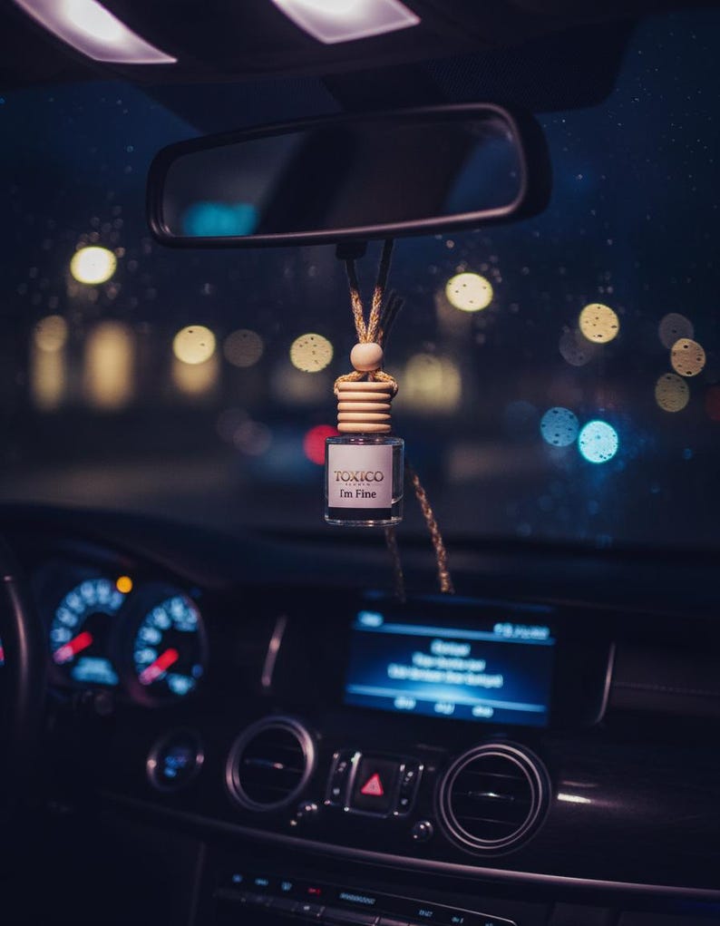 Puede incluir: Un ambientador de coche con el texto "TOXICO I'm Fine" cuelga del espejo retrovisor. Se ve el interior del coche, con el salpicadero, los indicadores y una pantalla iluminada en azul. El fondo muestra luces borrosas de la ciudad.