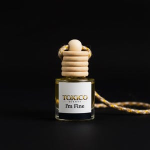 Puede incluir: Una peque&ntilde;a botella de vidrio transparente de ambientador Toxico Scents con el texto "I'm Fine" en negro y dorado. La botella tiene una tapa de madera y una cuerda decorativa. El fondo es negro.